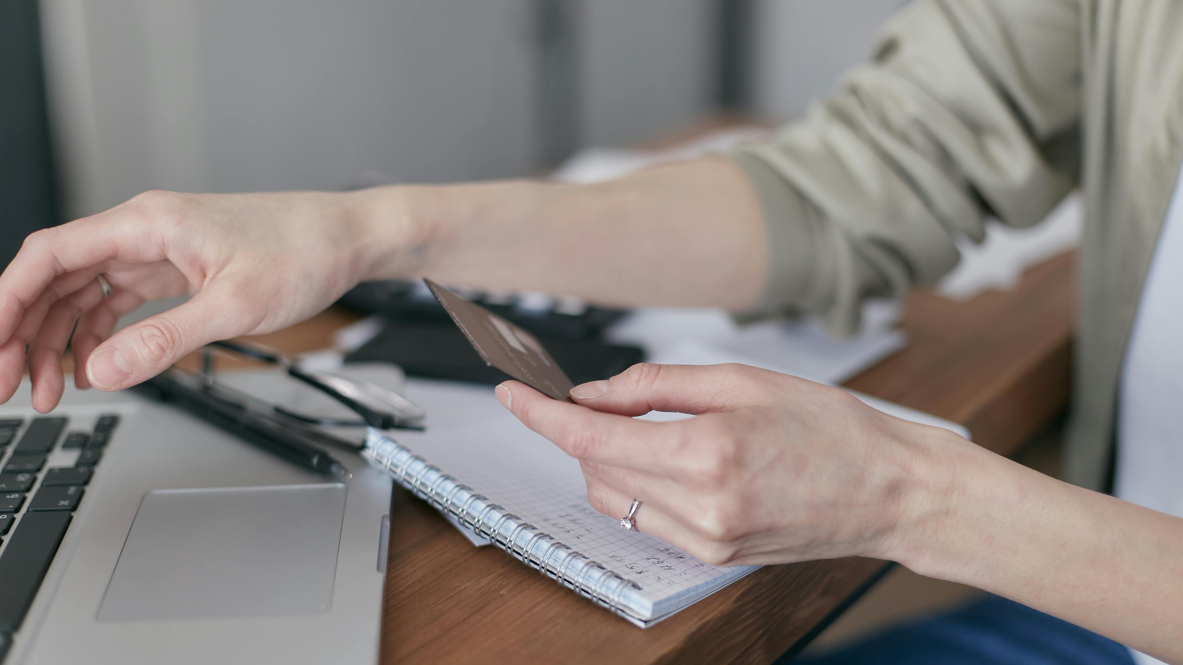 Hand reaching for a credit card next to an open laptop and notebook on a desk